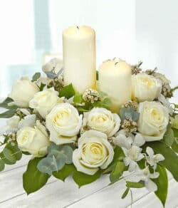 Classic Candle Arrangement featuring elegant white roses, orchids, and wax flowers with lush greenery, centred around a glowing white candle.