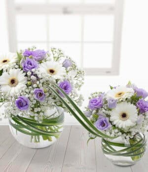 Bambino Flower Bouquet Featuring Lilac Lisianthus, White Germini, And Gypsophila In A Spherical Glass Vase, Linked To A Smaller Vase With Variegated China Grass.