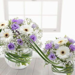Bambino Flower Bouquet featuring lilac Lisianthus, white Germini, and Gypsophila in a spherical glass vase, linked to a smaller vase with Variegated China Grass.