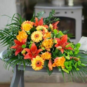 Amber Spray Arrangement Featuring Burnt Orange Asiatic Lilies, Orange Roses, Lime Green Spray Chrysanthemums, And Fresh Seasonal Foliage In A Teardrop Shape.